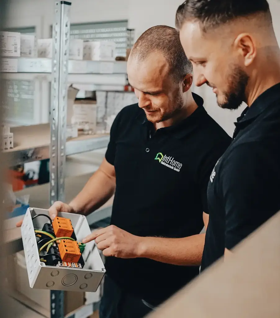 Zwei Mitarbeiter, Markus Steinbock und Markus Zink, betrachten konzentriert elektrische Bauteile in einem Verteilerkasten im Lager von IntHome.