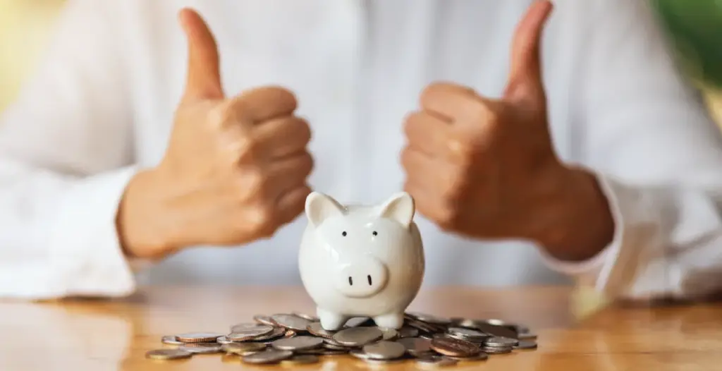 Eine Person zeigt beide Daumen hoch hinter einem Sparschwein und verdeutlicht Freude über die Ersparnis