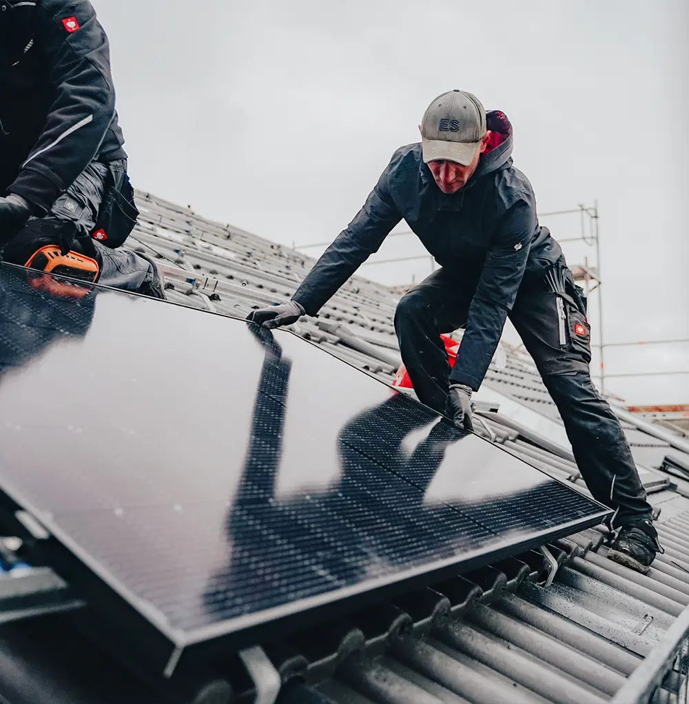 Monteure der IntHome Elektrotechnik GmbH installieren eine Photovoltaik-Anlage auf einem Dach. Sie sind bei der sorgfältigen Platzierung eines Solarmoduls zu sehen, gekleidet in Arbeitskleidung, konzentriert auf ihre Arbeit, um nachhaltige Energie zu gewährleisten.