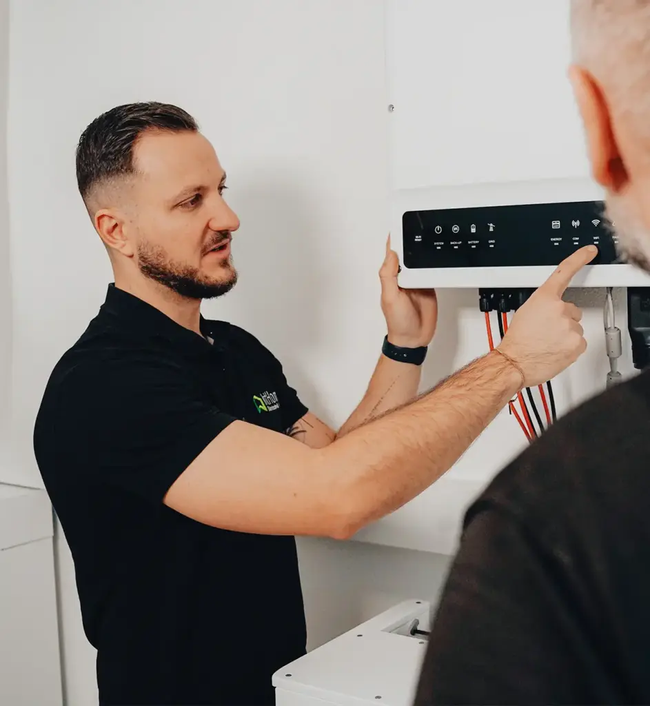 Ein Techniker von IntHome Elektrotechnik GmbH erklärt einem Kunden die Bedienung eines Solarwechselrichters. Er zeigt auf das Display und erläutert die verschiedenen Funktionen und Anzeigen. Diese Szene verdeutlicht den Kundenservice und die Fachkompetenz von IntHome bei der Installation und Wartung von Photovoltaikanlagen.