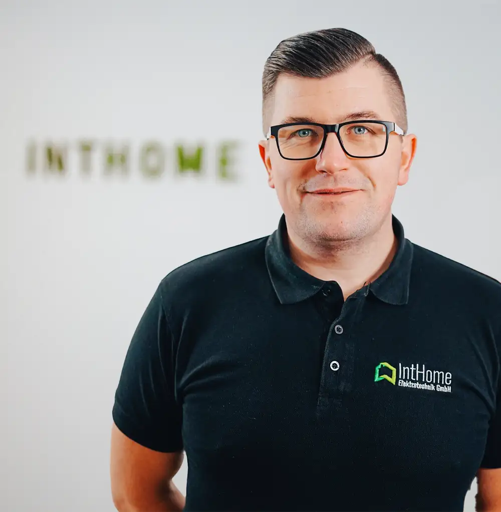 Piotr Wilkowski, Elektrohelfer bei IntHome Elektrotechnik GmbH, steht vor einer Wand mit dem Firmenlogo im Hintergrund. Er trägt ein schwarzes Polo-Shirt und schaut mit einem freundlichen und selbstbewussten Lächeln direkt in die Kamera.