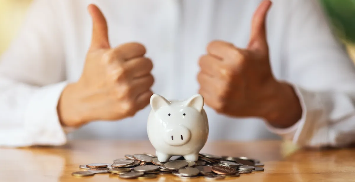 Eine Person zeigt beide Daumen hoch hinter einem Sparschwein und verdeutlicht Freude über die Ersparnis