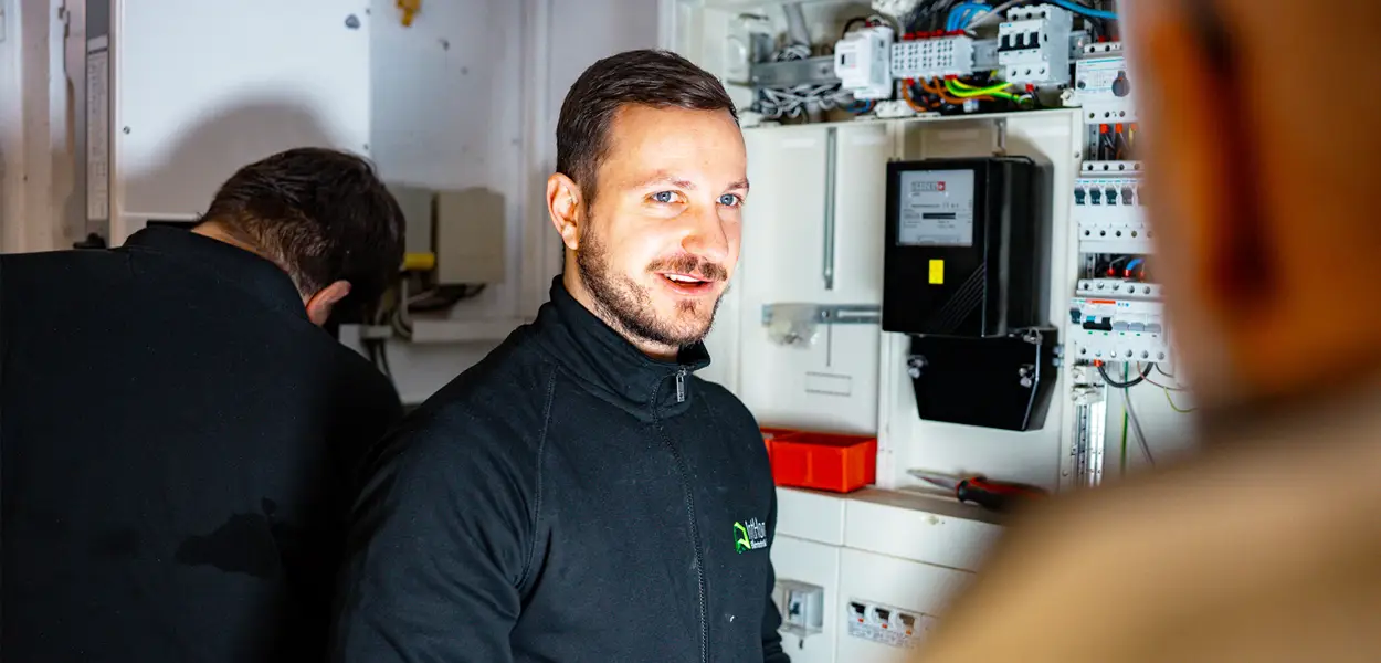 Markus Zink im Beratungsgespräch mit einem Kunden, zu sehen im Vordergrund eines Technikraums mit elektrischen Installationen.